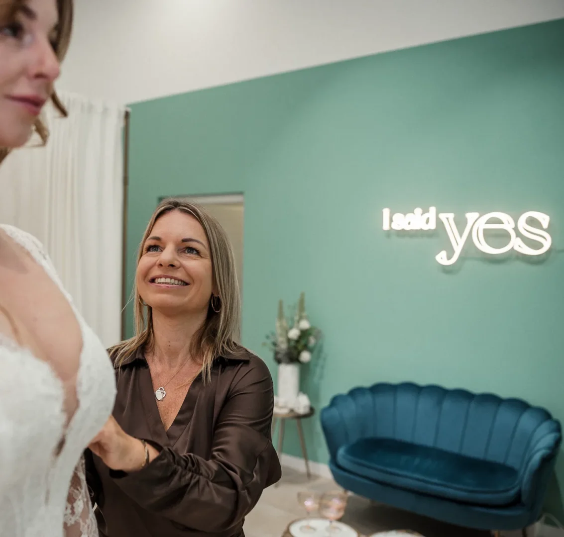 Ina von I said yes lacht mit einer Braut im Spitzenkleid, im Hintergrund der Neonschriftzug des Brautladens in Bad Rothenfelde